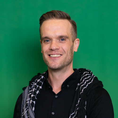 Portret van Bart tegen een donkergroene achtergrond, in zwart overhemd met korte mouwen en een zwarte keffiyeh met witte details over de schouders. Ik lach in de camera en heb mijn haren in een scheiding.